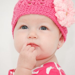 以下が含まれることがあります： ピンクの毛糸で編まれた帽子をかぶった赤ちゃん。帽子にはピンクの花が飾られています。赤ちゃんは上を見上げて、指を口に入れてます。