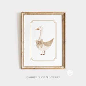 May include: A framed watercolour print featuring a goose with a bow. The goose has a white body, tan wings, and a long neck. The bow is a light tan colour. The print is in a wooden frame with a white background and a decorative border.