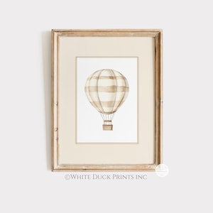 May include: A framed watercolour print featuring a hot air balloon. The balloon has beige and white stripes and a brown basket. The frame is light brown. The text "WHITE DUCK PRINTS INC" is at the bottom of the print.