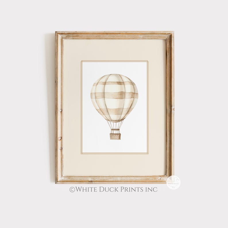 May include: A framed watercolour print featuring a hot air balloon. The balloon has beige and white stripes and a brown basket. The frame is light brown. The text "WHITE DUCK PRINTS INC" is at the bottom of the print.