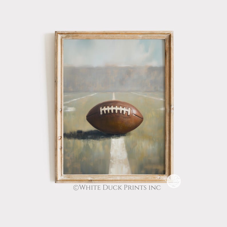 Peut inclure: Impression encadr&eacute;e d'un ballon de football marron sur un terrain. Le ballon repose sur une ligne blanche. L'arri&egrave;re-plan pr&eacute;sente un champ vert et un ciel brumeux. Le cadre est de couleur bois clair. Le texte "&copy;WHITE DUCK PRINTS INC" est en bas.