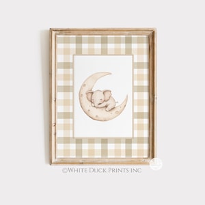 May include: A framed print with a watercolour illustration of a sleeping baby elephant on a crescent moon. The artwork is set against a white background and framed with a beige and white plaid pattern. The frame is a light brown colour. The text "WHITE DUCK PRINTS INC" is at the bottom.