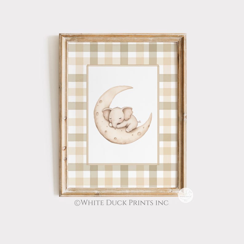May include: A framed print with a watercolour illustration of a sleeping baby elephant on a crescent moon. The artwork is set against a white background and framed with a beige and white plaid pattern. The frame is a light brown colour. The text "WHITE DUCK PRINTS INC" is at the bottom.