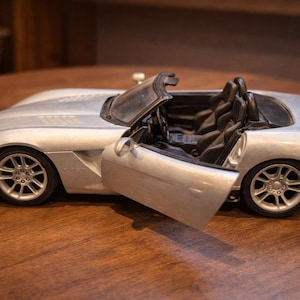 May include: Silver convertible model car with an open door, revealing black interior. The car has detailed wheels and a sleek design, sitting on a wooden surface. The car's headlights and taillights are visible.