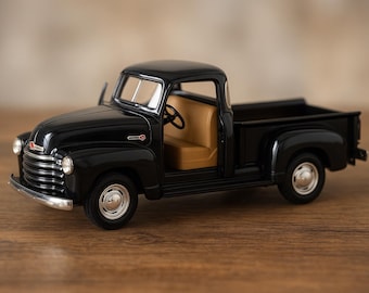May include: A black vintage pickup truck model with a tan interior. The truck has chrome accents on the front grill and bumper. The door is open, revealing the steering wheel and seats. The truck is sitting on a wooden surface.