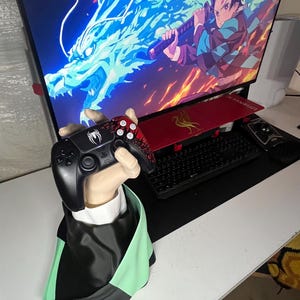May include: A black and red PlayStation 5 controller held by a statue of a hand. The controller has a spider design. The statue is wearing a black and green cape. A computer monitor displays an anime scene.