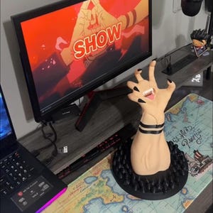 May include: A gaming setup featuring a computer monitor displaying the word "SHOW" in white against a red background. A hand-shaped figurine with black fingernails and a tongue is in the foreground. A laptop and keyboard are also visible.