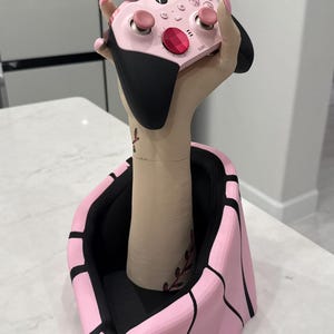 May include: A pink and black Xbox controller held by a decorative hand. The hand is part of a pink and black stand. The controller has pink buttons and a red center button. The stand is on a white surface.