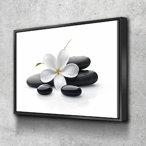 May include: A framed canvas print featuring a white flower and black spa stones. The flower has five petals and a yellow center. The stones are smooth and wet, with water droplets visible. The background is white, and the frame is black.