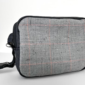 May include: A gray fanny pack with a plaid pattern in pink and gray. The bag has a black zipper, black trim, and a black adjustable strap with a buckle. The bag is rectangular and appears to be made of fabric.