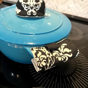 May include: A bright turquoise enamelled cast iron pot with a matching lid. The pot features a black and white damask patterned fabric handle cover and a matching fabric knob cover, both secured with small black fabric tabs for easy handling.