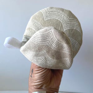 May include: A rounded, beige fabric oven mitt featuring a repeating white chevron or wave pattern. The mitt includes a small white fabric loop attached to the side for hanging, constructed from a textured, woven material with a curved shape.