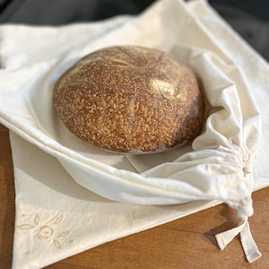 May include: A round loaf of bread with a textured, golden-brown crust rests inside a cream-colored cloth bread bag. The bag is tied at the top with a matching drawstring. The bag has a floral embroidery design on the corner.