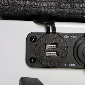 May include: A black charging station with two USB ports and a 12V outlet. The text "Charging Station" is printed on the station.