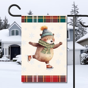 May include: A winter-themed garden flag with a brown bear wearing a blue and green hat and scarf, ice skating on a white background with snowflakes. The flag has a red and green plaid border.