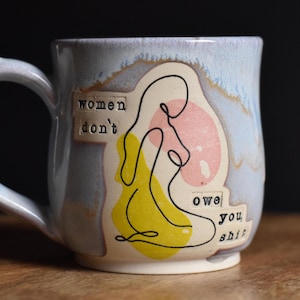 Può includere: Una tazza in ceramica con smalto azzurro chiaro e manico. La tazza presenta un disegno a tratto di una figura in giallo e nero, con sfondo rosa. Il testo sulla tazza recita "women don't owe you shit."