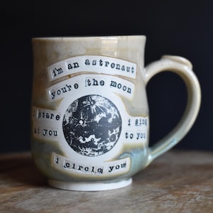 Könnte beinhalten: Ein Keramikbecher mit beiger und blaugrauer Glasur und einer zentralen schwarz-weißen Mondgrafik. Der Text lautet: I'm an astronaut, you're the moon, I stare at you, I sing to you, I circle you, in einer Schreibmaschinenschrift.