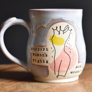 Puede incluir: Una taza de cerámica con un esmalte azul claro y marrón. La taza presenta un dibujo lineal negro de una figura con acentos amarillos y rosas. Las palabras "support women's rights" están impresas en la taza.