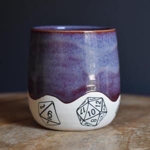 May include: A ceramic tumbler featuring a two-tone design with a glossy purple and blue glaze on the upper portion and a white base. The white section is decorated with black line art illustrations of various polyhedral gaming dice.