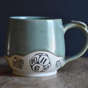 May include: A ceramic mug with a sage green glazed upper section and a white lower section. The white base features black ink illustrations of multi-sided dice, including a twenty-sided die with the numbers 10, 11, 6, and 5 clearly visible.