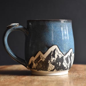 Puede incluir: Una taza de cerámica con una sección superior esmaltada en azul y una sección inferior blanca decorada con una ilustración en tinta negra de una cordillera. La taza tiene un asa curva con un pequeño acento circular en la parte superior.
