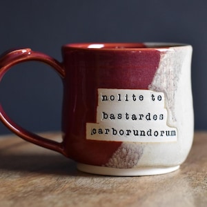 May include: A handmade ceramic mug with a burgundy handle and rim, and a white and burgundy body. The mug features the text "nolite te bastardes carborundorum" on a white label. The mug is on a wooden surface.