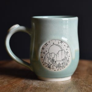 May include: A light blue ceramic mug with a black and white illustration of a desert scene with a tent, campfire, and cacti under a full moon and stars.