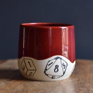 May include: A ceramic mug with a deep red glaze on the upper section and a cream-coloured base. The base features black illustrations of dice with numbers. The mug is cylindrical and sits on a wooden surface.