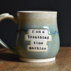 May include: A handmade ceramic mug with a unique design. The mug features a two-tone glaze in shades of green and blue, with a white base and handle. The text "I am a breathing time machine" is printed on the front.