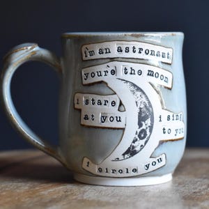 Könnte beinhalten: Ein hellblauer Keramikbecher mit geschwungenem Henkel, verziert mit einer weißen Mondsichel und dem schwarzen Text: I'm an astronaut, you're the moon, I stare at you, I sing to you, I circle you. Der Becher hat eine glatte, glasierte Oberfläche.