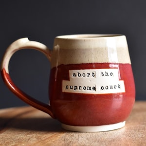 Puede incluir: Una taza de cerámica roja y blanca con el texto "abort the supreme court" en el lateral.