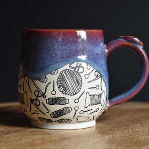 May include: A handmade ceramic mug with a blue and burgundy glaze. The mug features a white base with black line drawings of sewing notions, including spools of thread, needles, and scissors. The handle is a matching burgundy color.