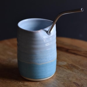 May include: A handcrafted ceramic tumbler in shades of light blue and teal, with a stainless steel straw. The cup is placed on a wooden surface. The tumbler is ideal for cold drinks.