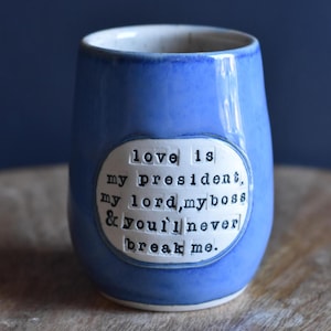 May include: A blue ceramic mug with a white oval sticker that reads "love is my president, my lord, my boss & you'll never break me."