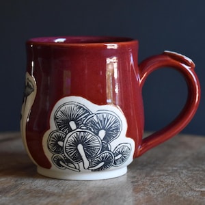 Può includere: Una tazza in ceramica rosso lucido con un inserto bianco che presenta un'illustrazione a inchiostro nero di un gruppo di funghi. La tazza ha un corpo arrotondato, un manico robusto e una finitura liscia con una stampa botanica dettagliata.