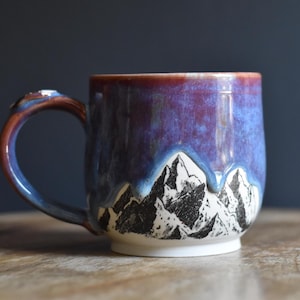 Könnte beinhalten: Ein Keramikbecher mit einer handgezeichneten schwarz-weißen Gebirgsillustration am unteren Rand. Der obere Teil des Bechers ist mit einer leuchtenden Glasur in Violett und Blau überzogen, die über die Bergspitzen nach unten verläuft.