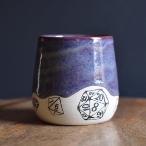 May include: A handmade ceramic tumbler with a purple and blue glaze. The bottom half is white with black illustrations of polyhedral dice. The tumbler sits on a wooden surface.