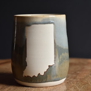 May include: A ceramic tumbler featuring a white outline of Indiana. The mug has a gradient of green, blue, and tan hues. The mug is set on a wooden surface.