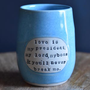 May include: A light blue ceramic mug with a white oval sticker that reads "love is my president, my lord, my boss & you'll never break me."