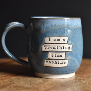 May include: A blue ceramic mug with a speckled glaze and a handle on the left side. The front features four white rectangular clay plaques with the black stamped text: i am a breathing time machine. The base of the mug is a solid white color.