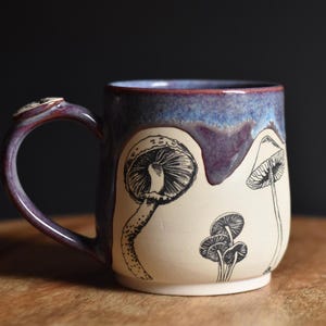 Può includere: Una tazza in ceramica con corpo color crema e illustrazioni a inchiostro nero di vari funghi. La parte superiore della tazza e il manico sono rifiniti con uno smalto lucido e maculato in tonalità viola e blu scuro che cola verso il basso.