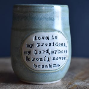 May include: A light green ceramic mug with a white oval sticker that reads "love is my president, my lord, my boss & you'll never break me."