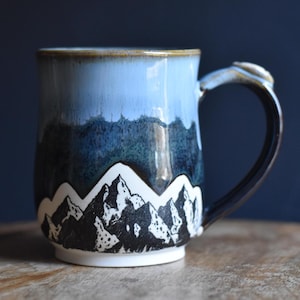 May include: A ceramic mug featuring a hand-painted mountain range scene at the base in black and white, transitioning into a deep blue glaze in the middle and a lighter sky blue glaze at the rim, complete with a sturdy curved handle.