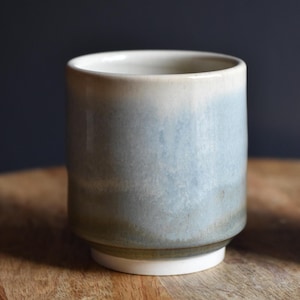 Puede incluir: Un vaso de cerámica hecho a mano con un degradado de color. El borde superior es blanco crema, pasando a azul claro y luego a un verde apagado cerca de la base. El vaso se asienta sobre una base blanca, ideal para bebidas. (aproximadamente 10 cm de alto)