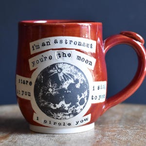 May include: A glossy burnt-orange ceramic mug featuring a central black and white graphic of the moon. The text reads: I'm an astronaut you're the moon I stare at you I sing to you I circle you. The mug has a curved handle and a white base.
