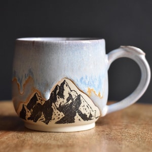 May include: A ceramic mug featuring a hand-drawn black mountain range illustration against a cream-colored base. The upper portion of the mug is glazed in a light blue with drips of tan and white, creating a layered effect around the handle.