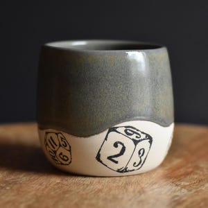 May include: A ceramic mug featuring a two-tone design with a smooth, dark olive-green glaze on the upper portion and a matte off-white base. The lower section is decorated with black ink illustrations of various polyhedral gaming dice.