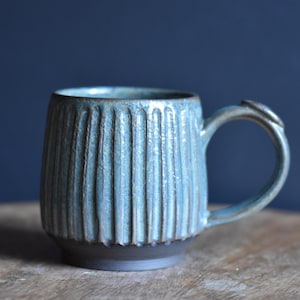 Op de afbeelding: Een handgemaakte keramische mok met een textuur, verticaal geribbeld ontwerp in tinten blauw en grijs. De mok heeft een donkere basis en een gebogen handvat. De mok is ontworpen voor warme dranken.