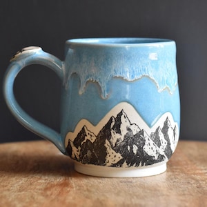 May include: A ceramic mug featuring a light blue glaze that drips down over a white base. The lower portion of the mug displays a detailed black and white ink-style illustration of a mountain range with jagged peaks and textured rocky slopes.
