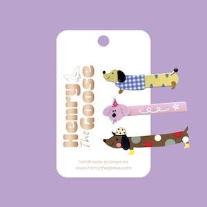 May include: Set of four colorful wooden hair clips featuring dachshund dog designs. The clips are decorated with polka dots, stripes, and a party hat. The clips are handmade and feature the text "Henry The Goose" and "handmade accessories www.henrythegoose.com".